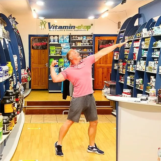Marcin, the owner, striking a pose in the middle of Vitamin Shop with shelves of supplements behind him